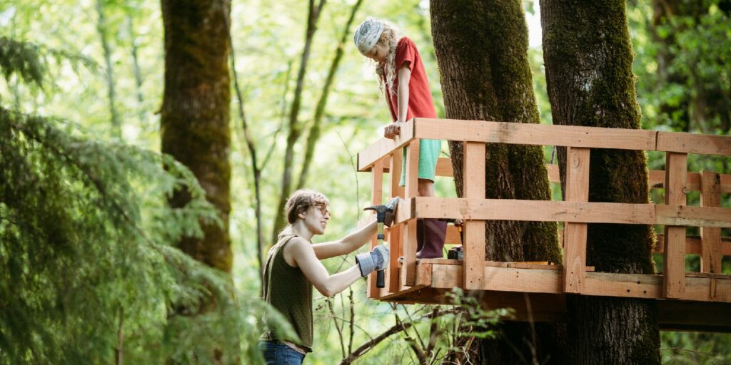A woman and her child work together on the railings of a tree house in a large maple in their wooded back yard