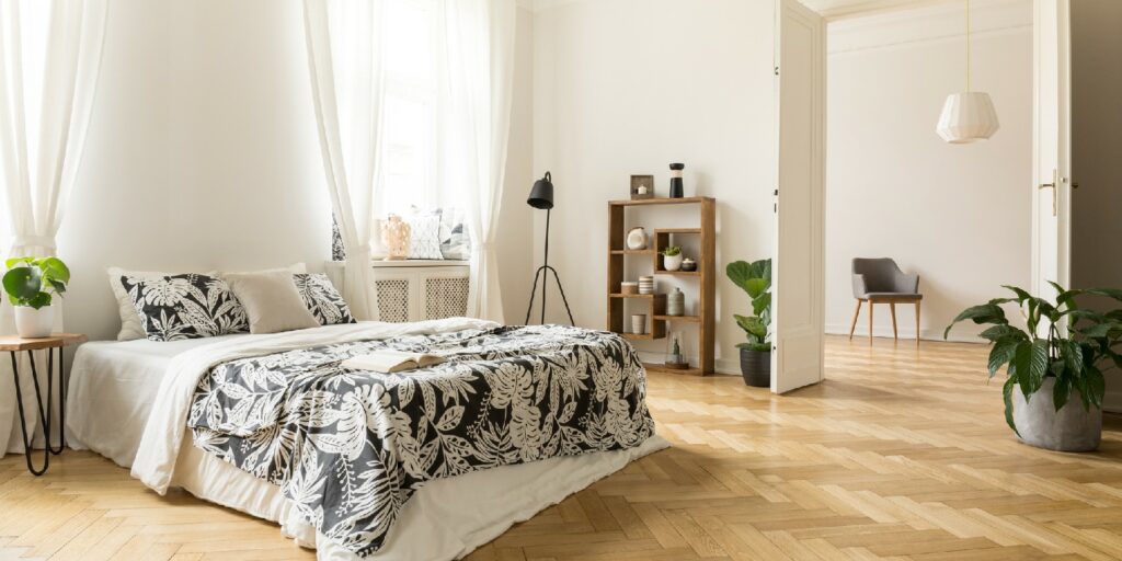 Stylish apartment interior with white walls and herringbone wooden floor. A view from a bedroom with a big bed to another room with an armchair. Real photo.